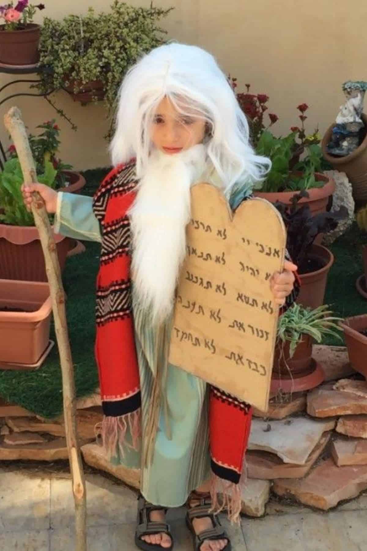Child in a costume with a white wig, fake beard, robe, and holding a staff and a cardboard tablet with Hebrew writing, standing in a garden with potted plants.