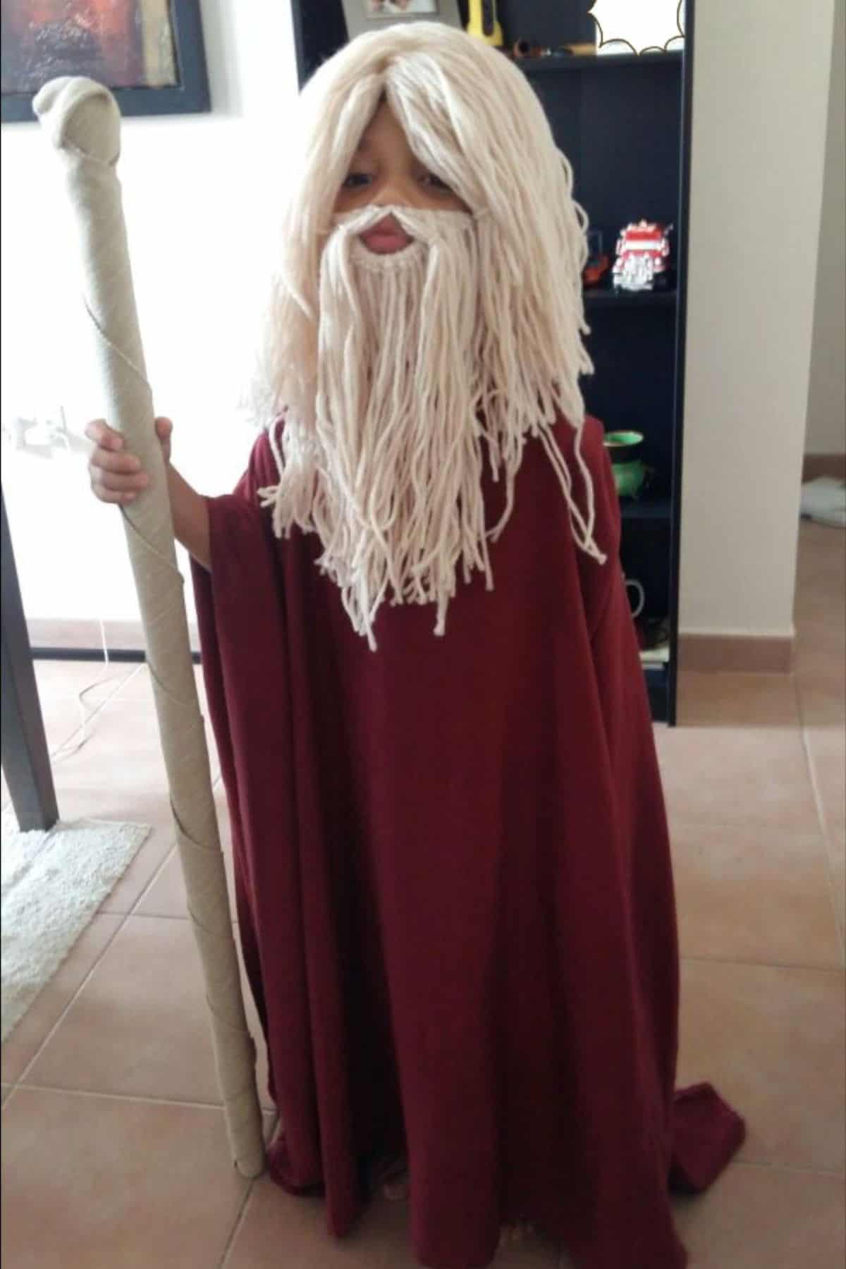 A child dressed as a wizard, wearing a maroon robe, a fake white beard made of yarn, and holding a tall staff, stands indoors on a tiled floor.