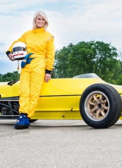 Woman in a yellow racing suit stands beside a yellow open-wheel race car on a paved surface with green trees in the background.