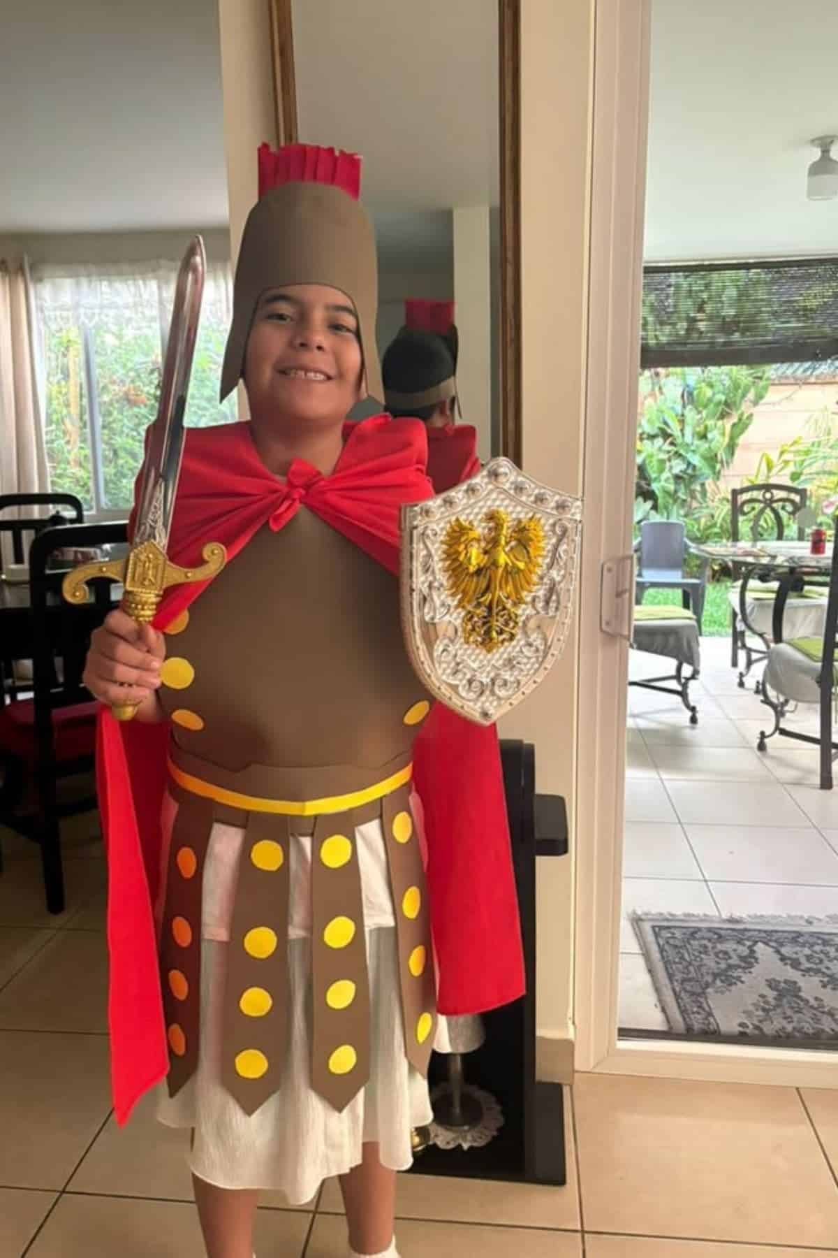 A child stands indoors dressed in a Roman soldier costume, holding a toy sword and a decorative shield, and smiling at the camera.