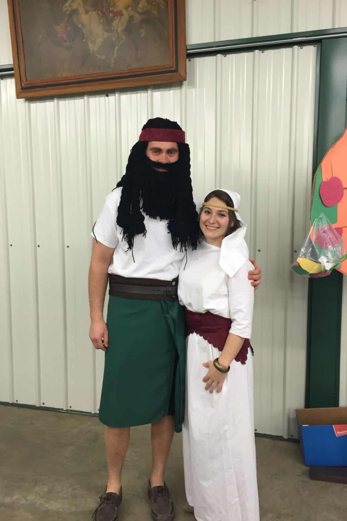 Two people in costume stand indoors. One wears a fake black beard, headband, green skirt, and belt; the other wears a white outfit with a headscarf and maroon sash.