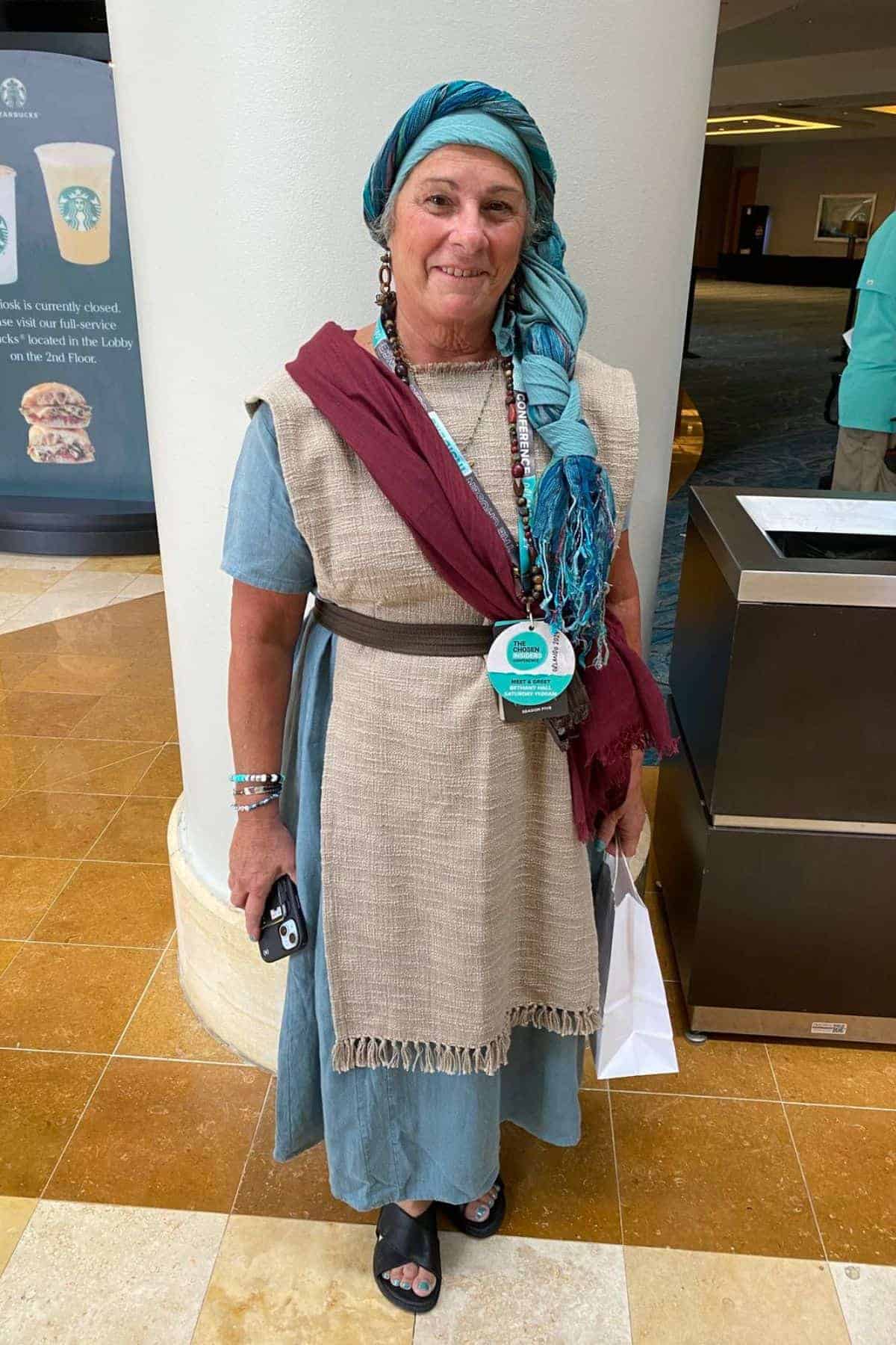 A woman wearing a blue dress, beige tunic, and blue headscarf stands indoors holding a white bag and a phone, with a badge around her neck.