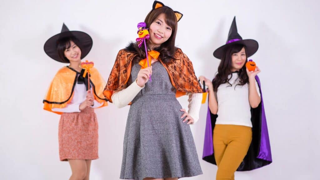 Three women in Halloween costumes pose against a white background; one wears cat ears, the others wear witch hats and capes, and all hold Halloween-themed props.
