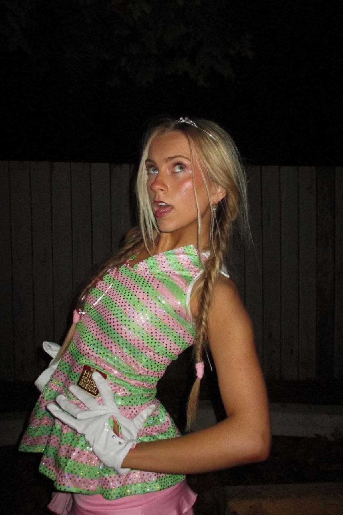 Person in a pink and green sequin outfit with braided hair poses with white gloves at night in front of a wooden fence.