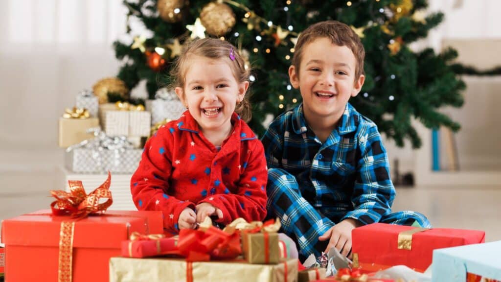 Two young children in pajamas sit smiling in front of a decorated Christmas tree, surrounded by wrapped gifts.