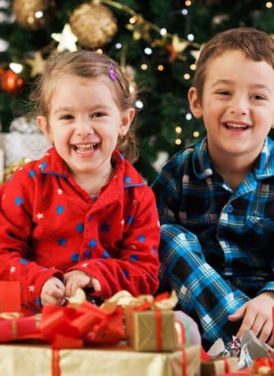 Two young children in pajamas sit smiling in front of a decorated Christmas tree, surrounded by wrapped gifts.