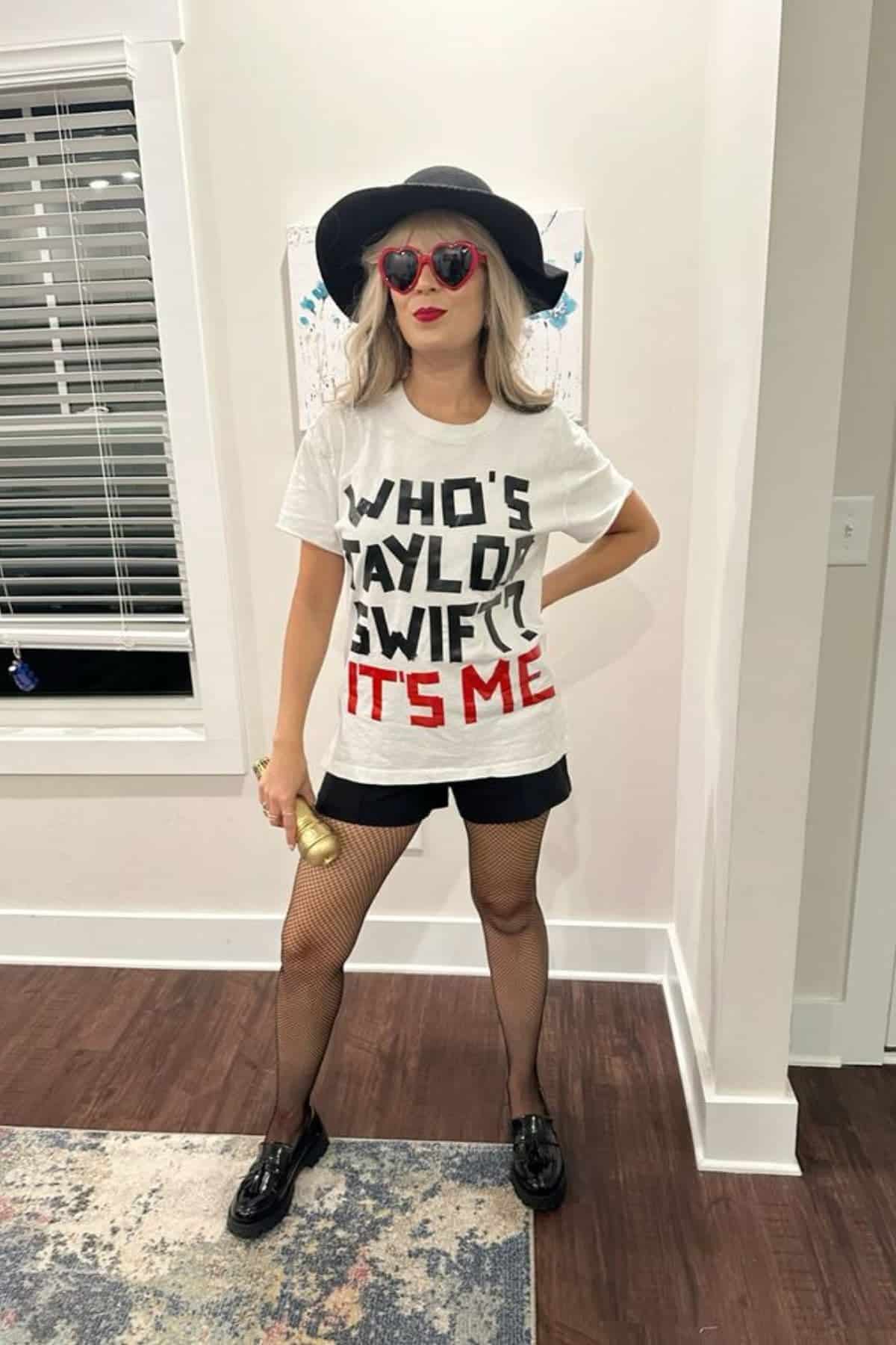 Person wearing a “Who’s Taylor Swift? It’s Me” shirt, black hat, red heart sunglasses, black shorts, fishnet stockings, and holding a gold microphone, standing indoors.