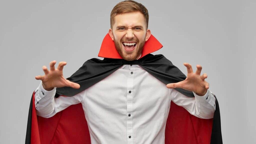 A man in a white shirt and black-and-red vampire cape poses with hands raised and mouth open, as if pretending to be a vampire, against a plain gray background.