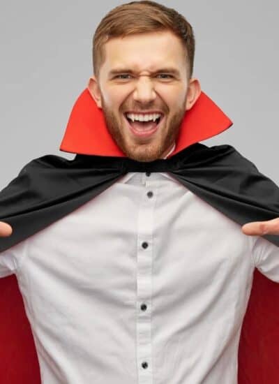 A man in a white shirt and black-and-red vampire cape poses with hands raised and mouth open, as if pretending to be a vampire, against a plain gray background.