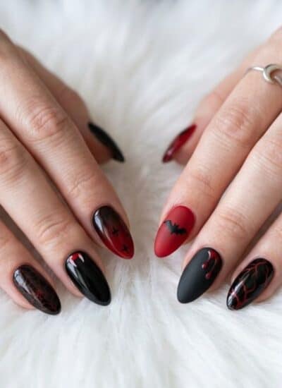 Hands with long, almond-shaped nails painted in black and red designs rest on a white, fluffy surface. The person wears two simple silver rings.