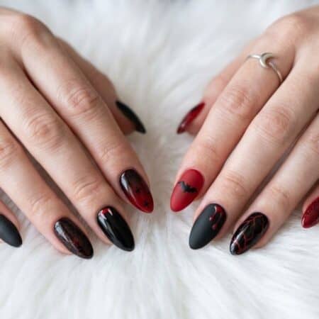 Hands with long, almond-shaped nails painted in black and red designs rest on a white, fluffy surface. The person wears two simple silver rings.