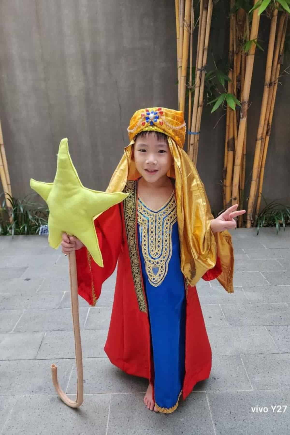 Child in a red and blue costume with gold headpiece holds a yellow star wand and stands barefoot on gray tiles near bamboo.