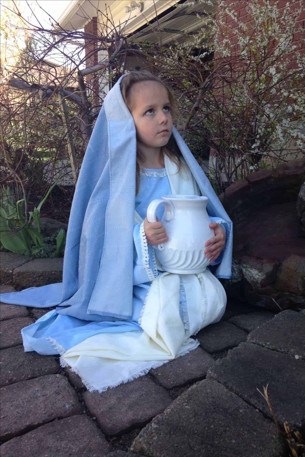 Young girl dressed in a blue and white robe sits on a stone path, holding a large white pitcher, with bare branches and a brick wall in the background.