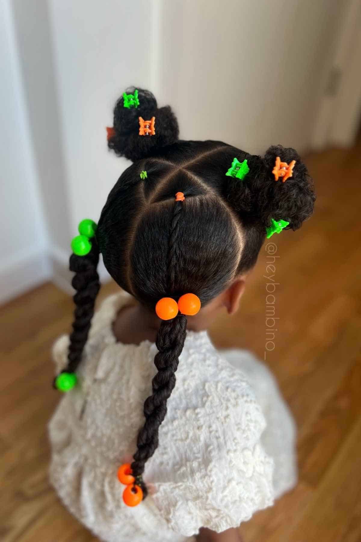 Child’s hairstyle with two top buns and two braids, sectioned into triangles and decorated with bright green and orange mini clips and beads.