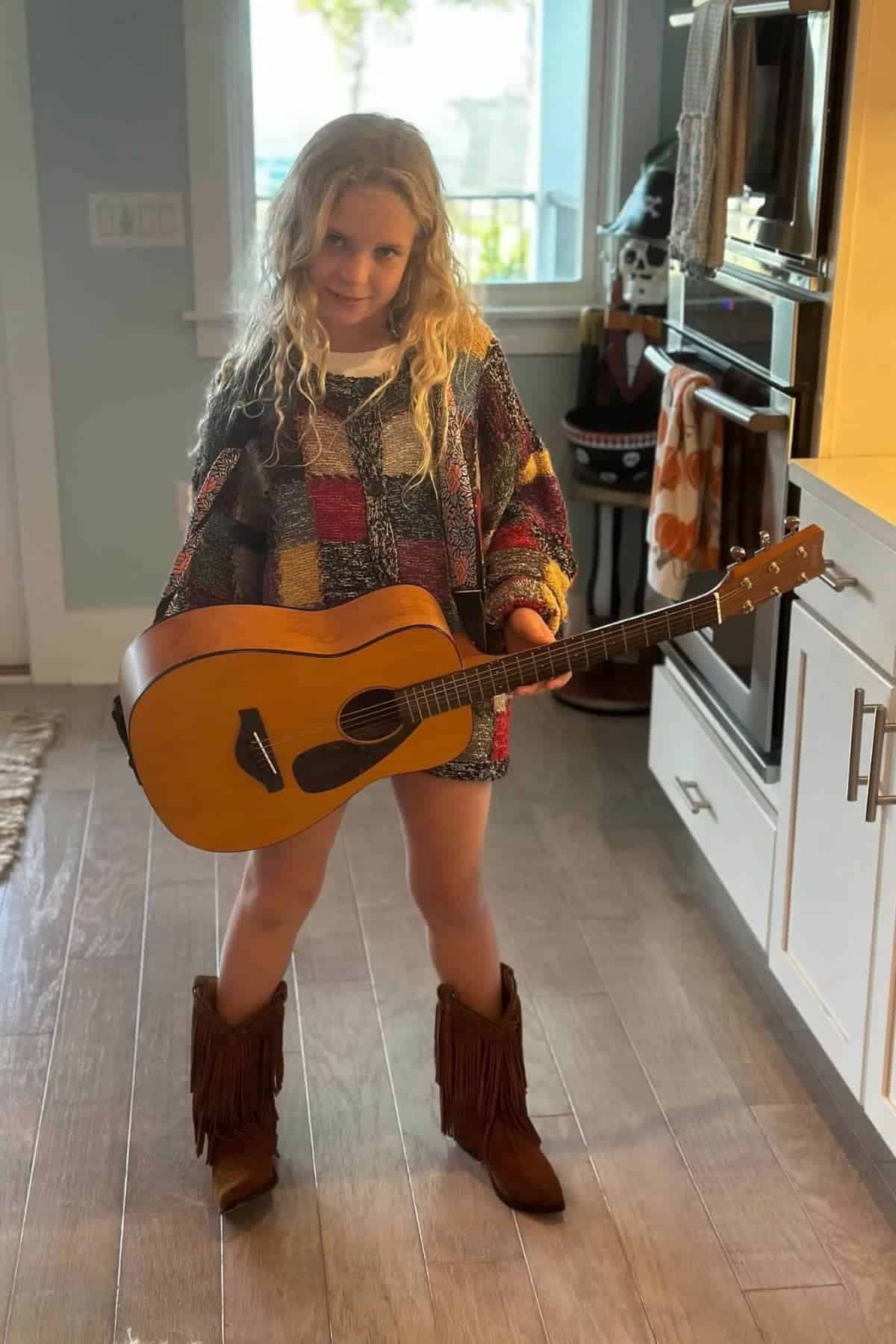 Young girl with wavy blonde hair holds a guitar and stands indoors on wood flooring, wearing a patchwork sweater and brown fringe boots.