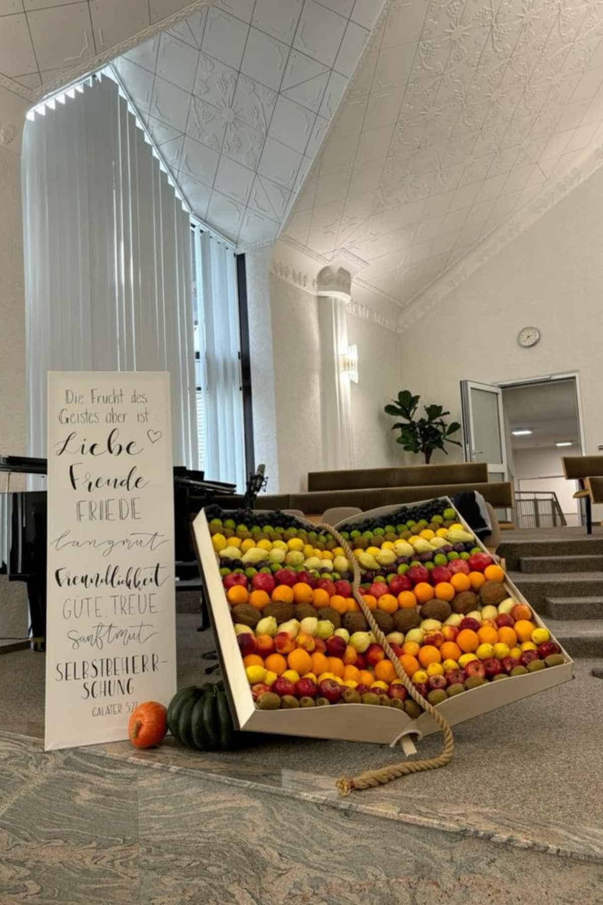 A sign listing the fruits of the spirit in German next to an open, book-shaped crate filled with rows of assorted fruits.