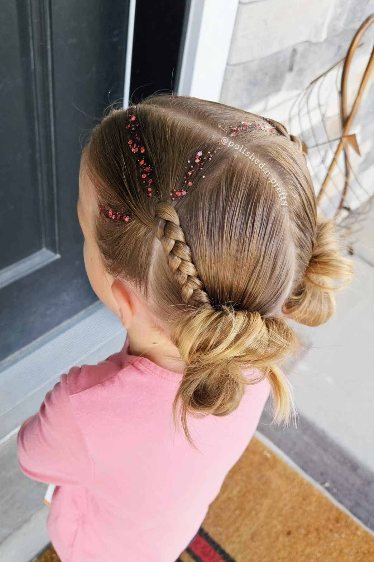 Child with a braided hairstyle featuring red glitter along the part and two small buns.