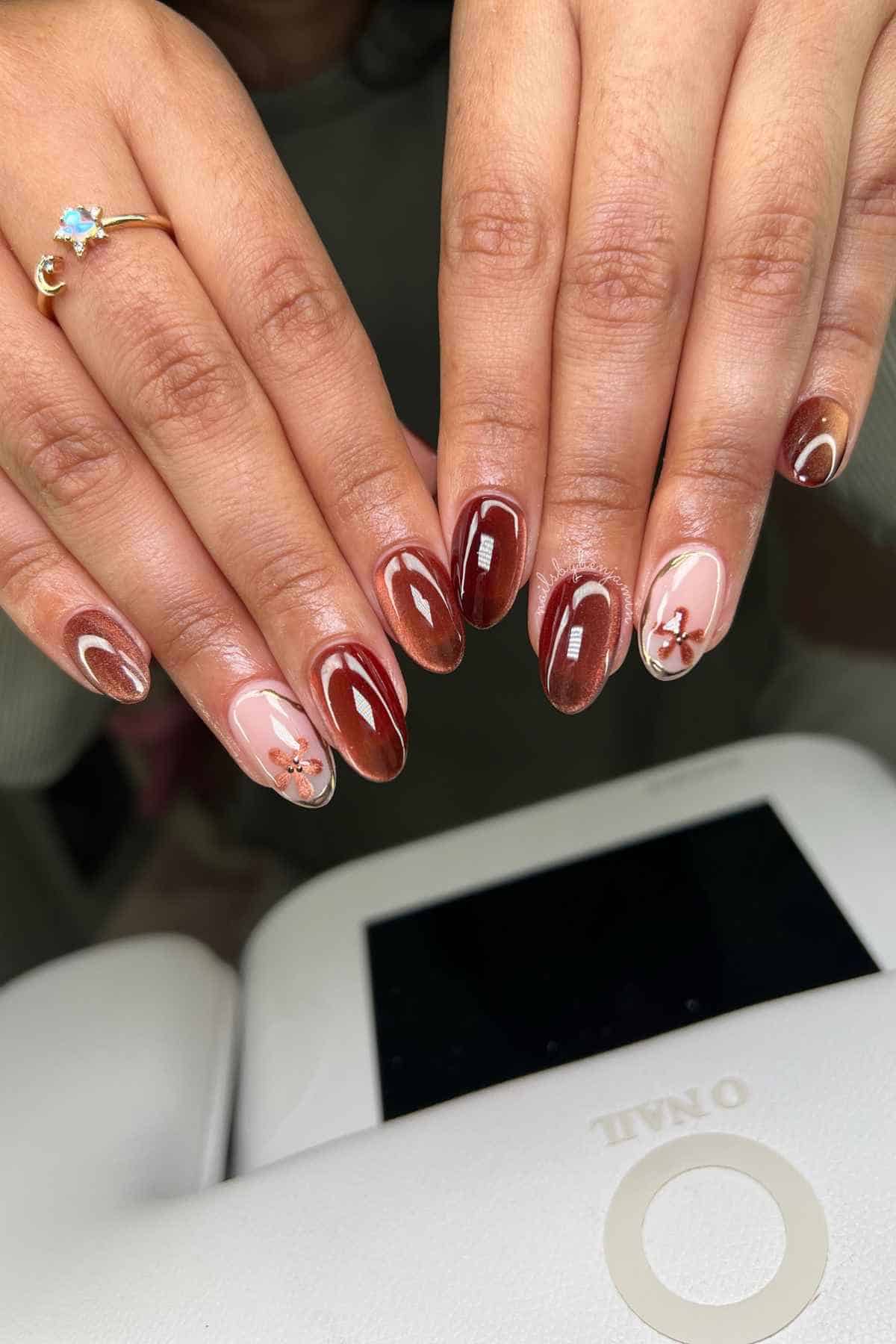 Hands with almond-shaped nails featuring a mix of glossy brown cat-eye polish and light pink accents with small flower decals.