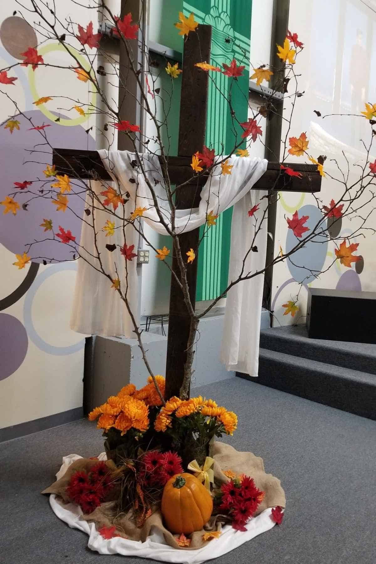 A wooden cross draped with white cloth, decorated with autumn leaves, orange flowers, and a pumpkin at the base.