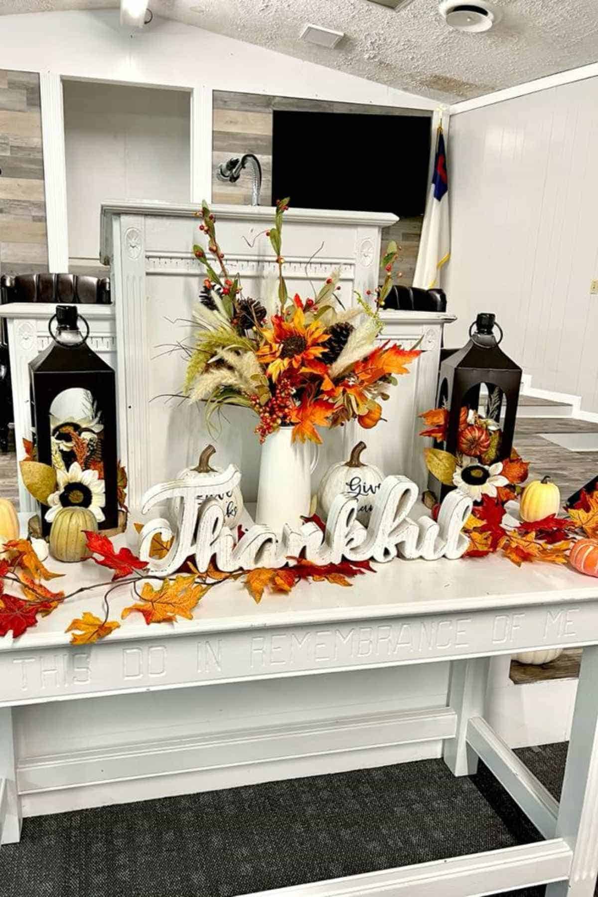 White communion table decorated with autumn leaves, pumpkins, lanterns, and a centerpiece vase with the word “Thankful” displayed in front.
