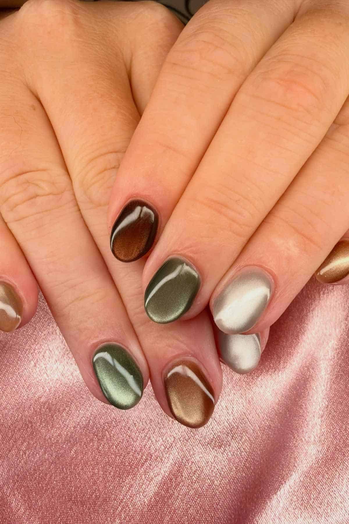 Hands showing almond-shaped nails painted with multi-toned, metallic cat-eye polish in shades of brown, green, and silver.