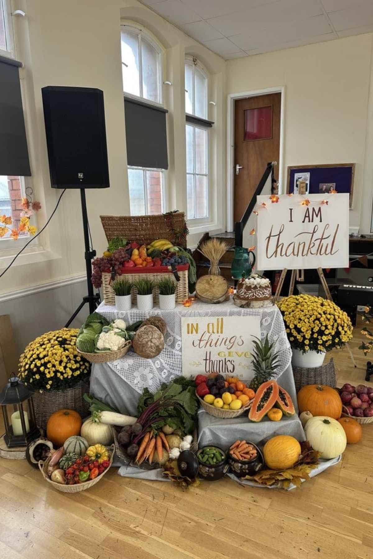 A harvest display featuring baskets of vegetables and fruit, two “thankful” themed signs, and potted yellow chrysanthemums on a table covered by a white lace cloth.
