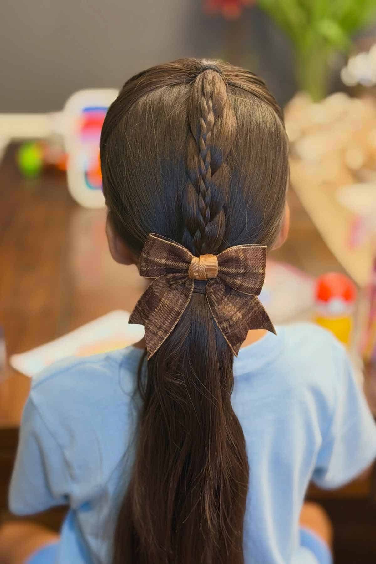 A child’s hair styled in a center braid leading into a ponytail, secured with a brown plaid bow.