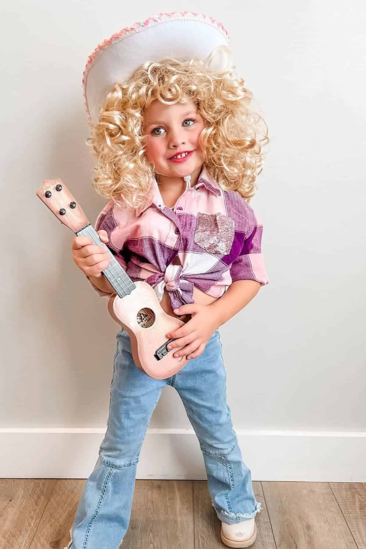 Child in curly blonde wig, white cowboy hat, plaid shirt, and jeans holds a small pink guitar, standing against a plain white wall.