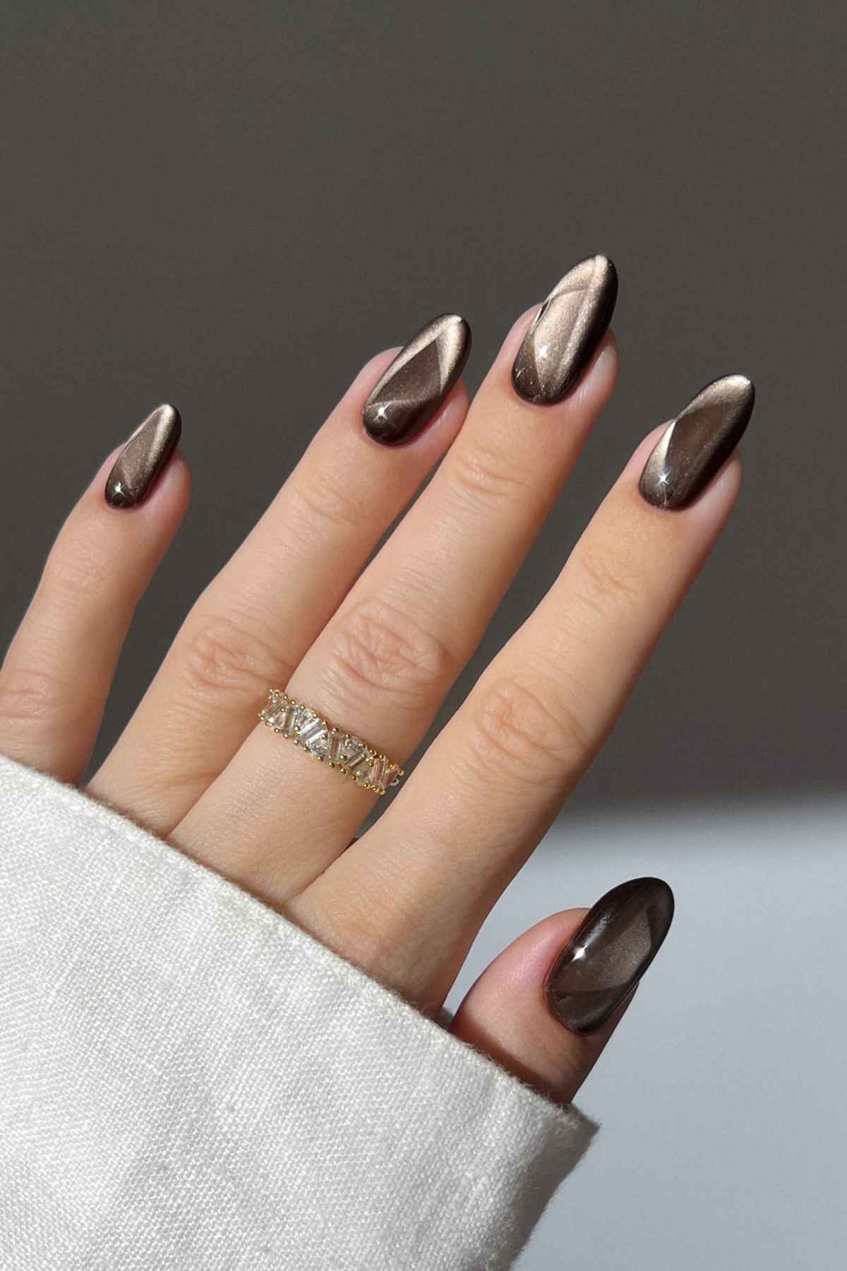 A hand with almond-shaped nails painted in a dark, reflective magnetic polish, wearing a thin gold band with clear stones.