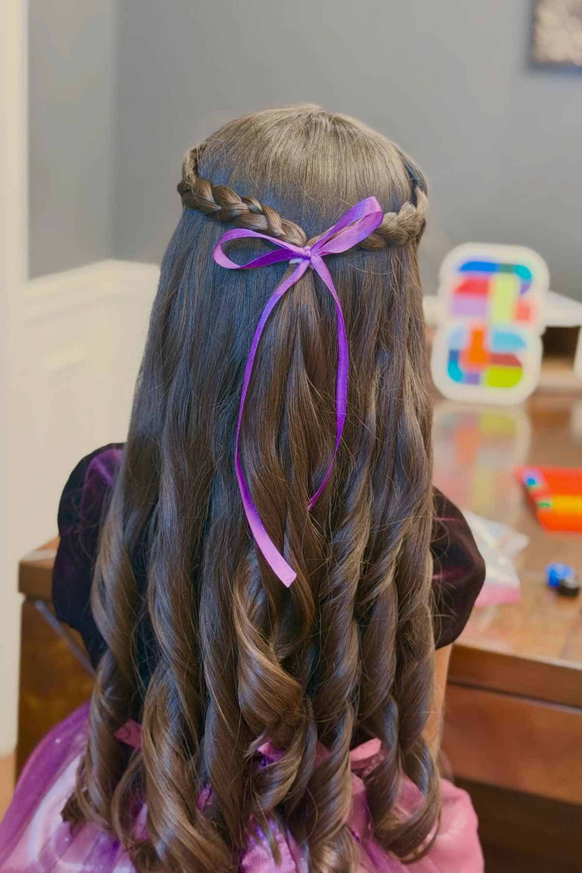 Long, wavy brown hair styled with a braided crown and a purple ribbon tied at the back.