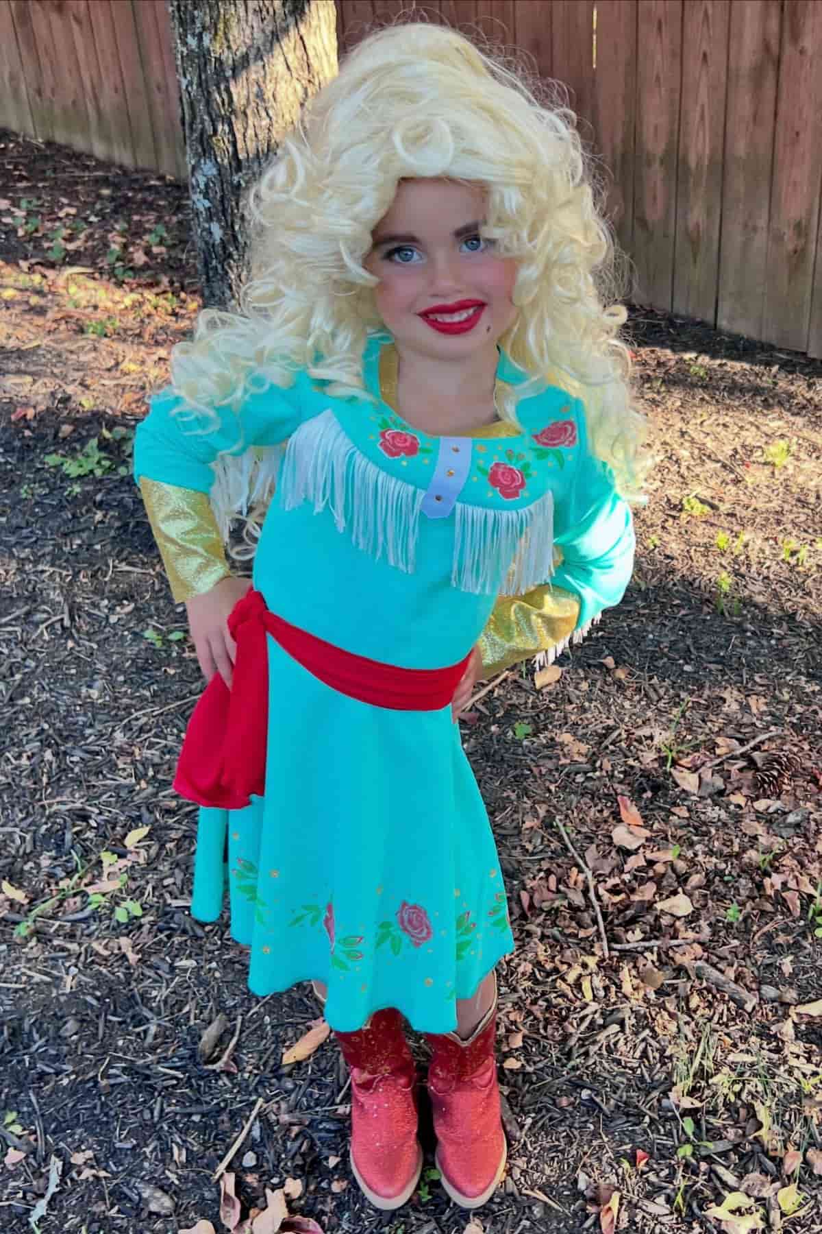 Young child outdoors wearing a long blonde wig, teal dress with floral embroidery, red sash, gold cuffs, and red cowboy boots, standing on dirt and leaves near a wooden fence.