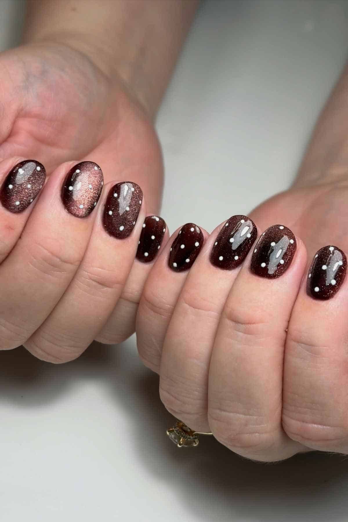 Dark brown magnetic cat-eye nail polish with white polka dots on rounded fingernails.