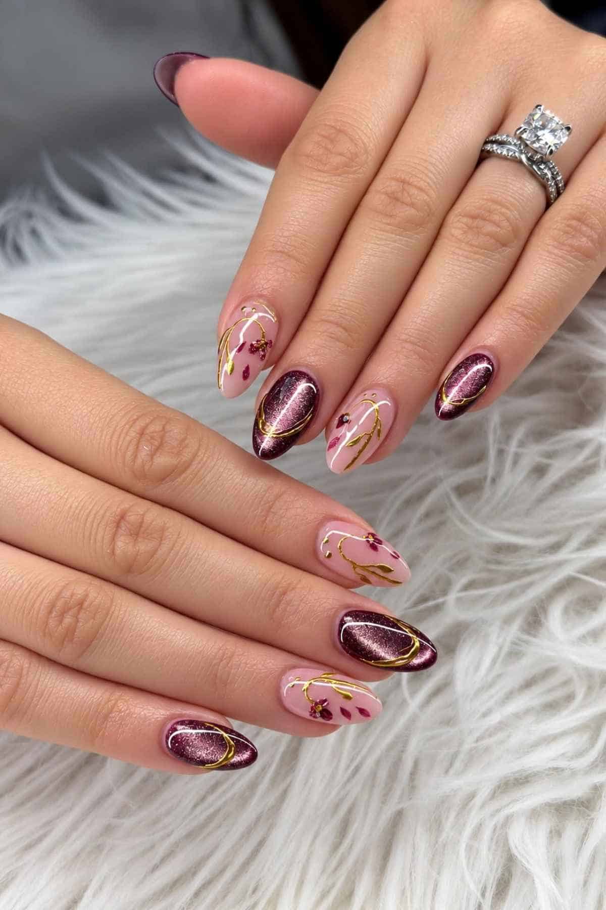 Manicured almond-shaped nails featuring a mix of dark plum cat-eye polish and light pink nails with gold floral decals.