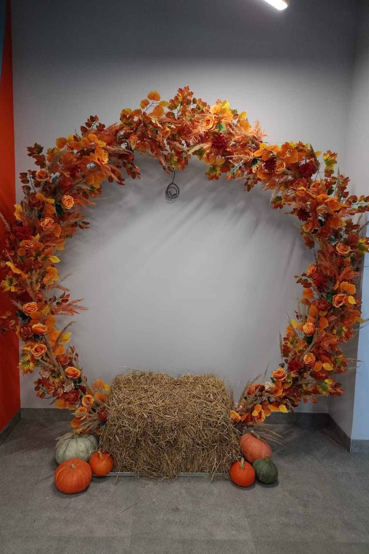 A circular arch decorated with autumn leaves and flowers, framing a square bale of hay on the floor with several pumpkins nearby.