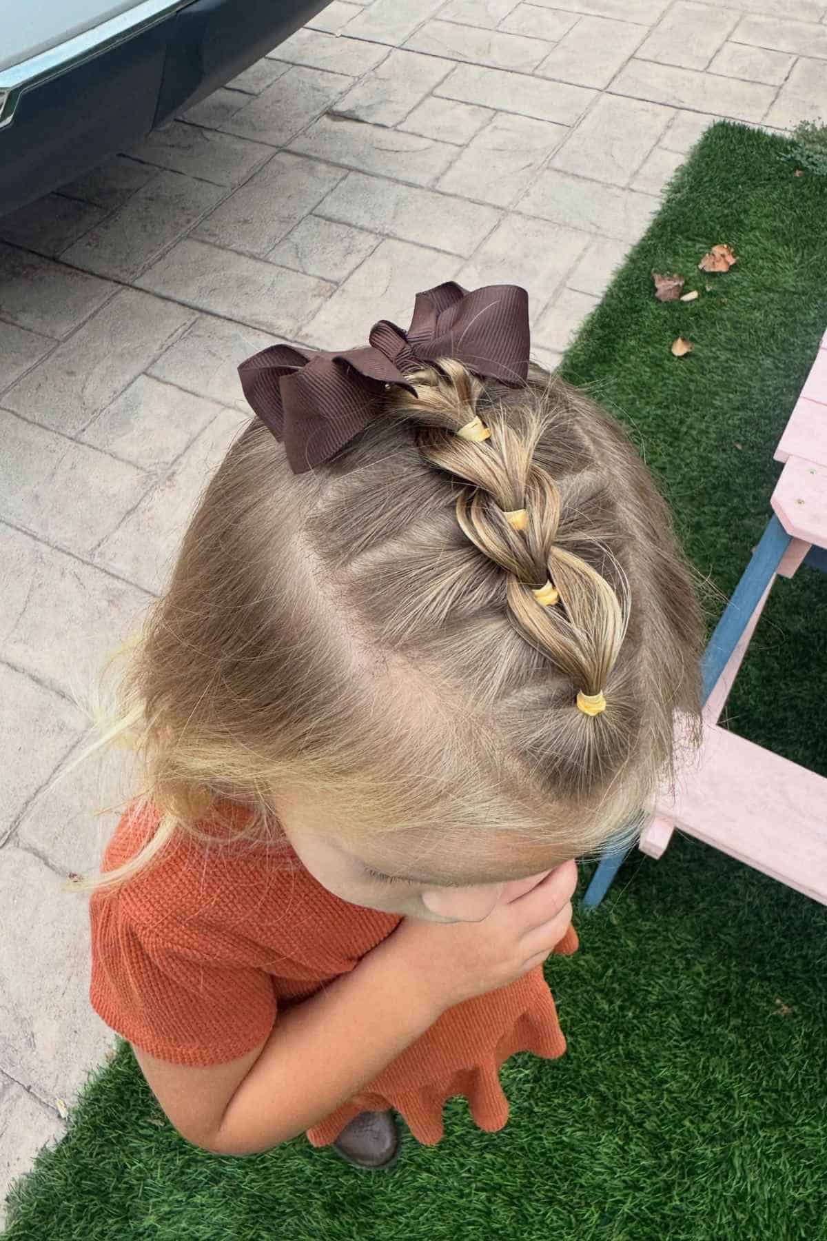 A young child with a brown hair bow and a French braid secured with small yellow elastics.