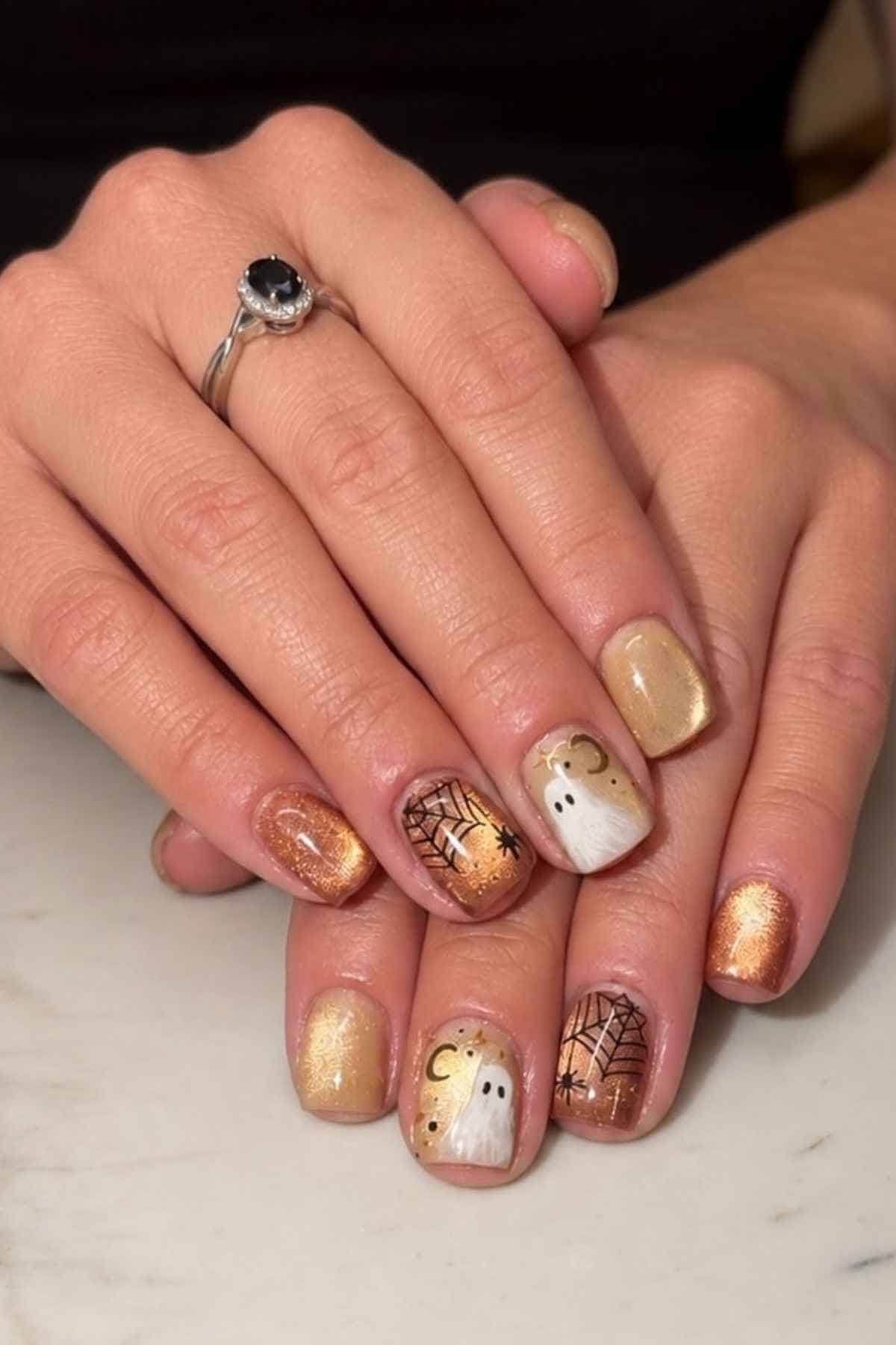 Hands showing a Halloween-themed manicure with gold shimmer polish, spiderweb art, and small ghost designs.