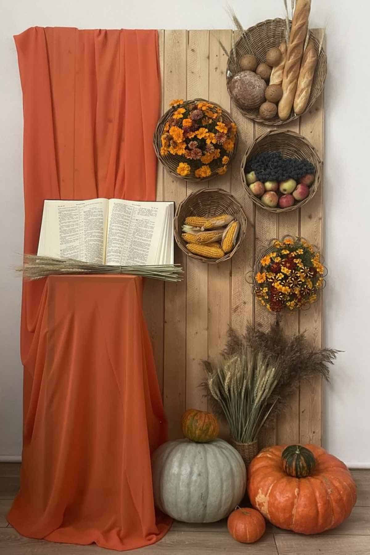 An open book rests on a pedestal draped in orange fabric, beside a wooden wall adorned with baskets of produce and flowers, and pumpkins on the floor.