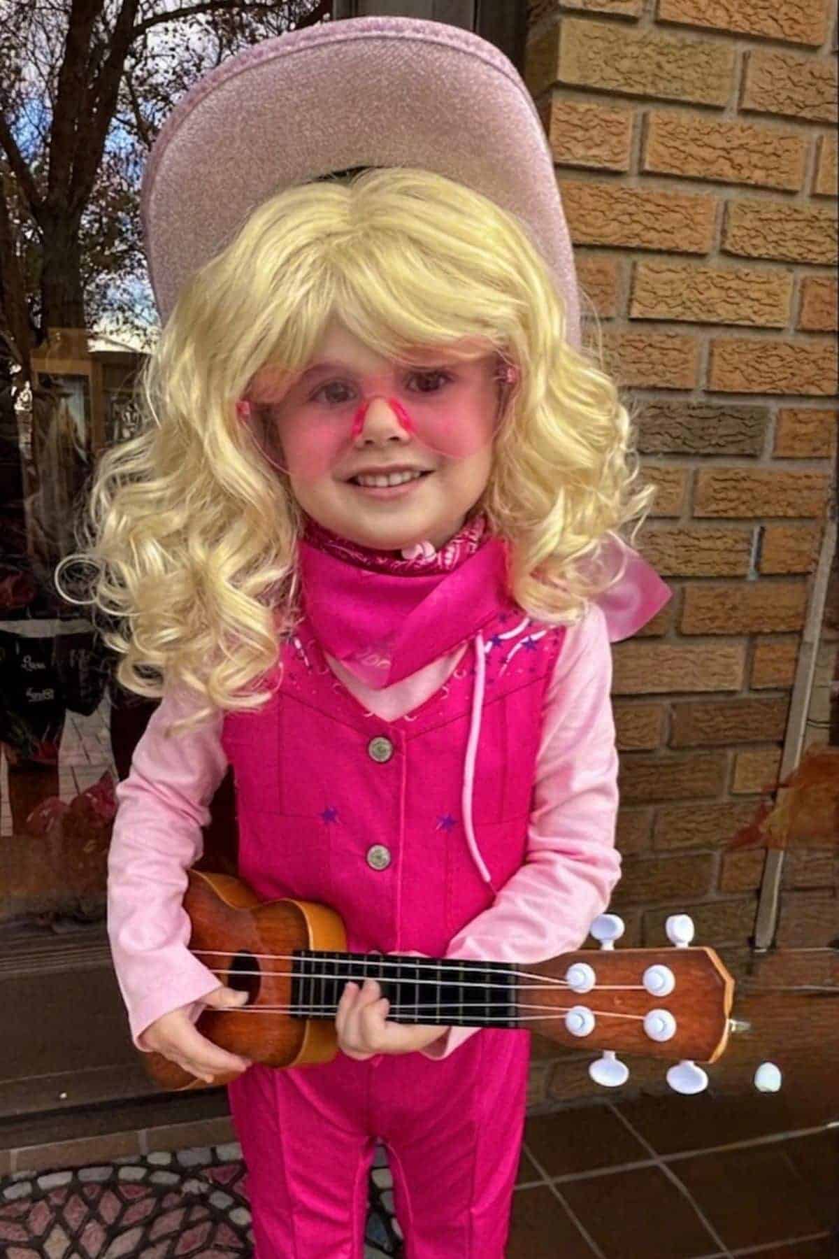 Child dressed in a pink cowboy outfit, with a blonde wig, pink hat, pink sunglasses, and scarf, holding a small ukulele, standing in front of a brick wall.