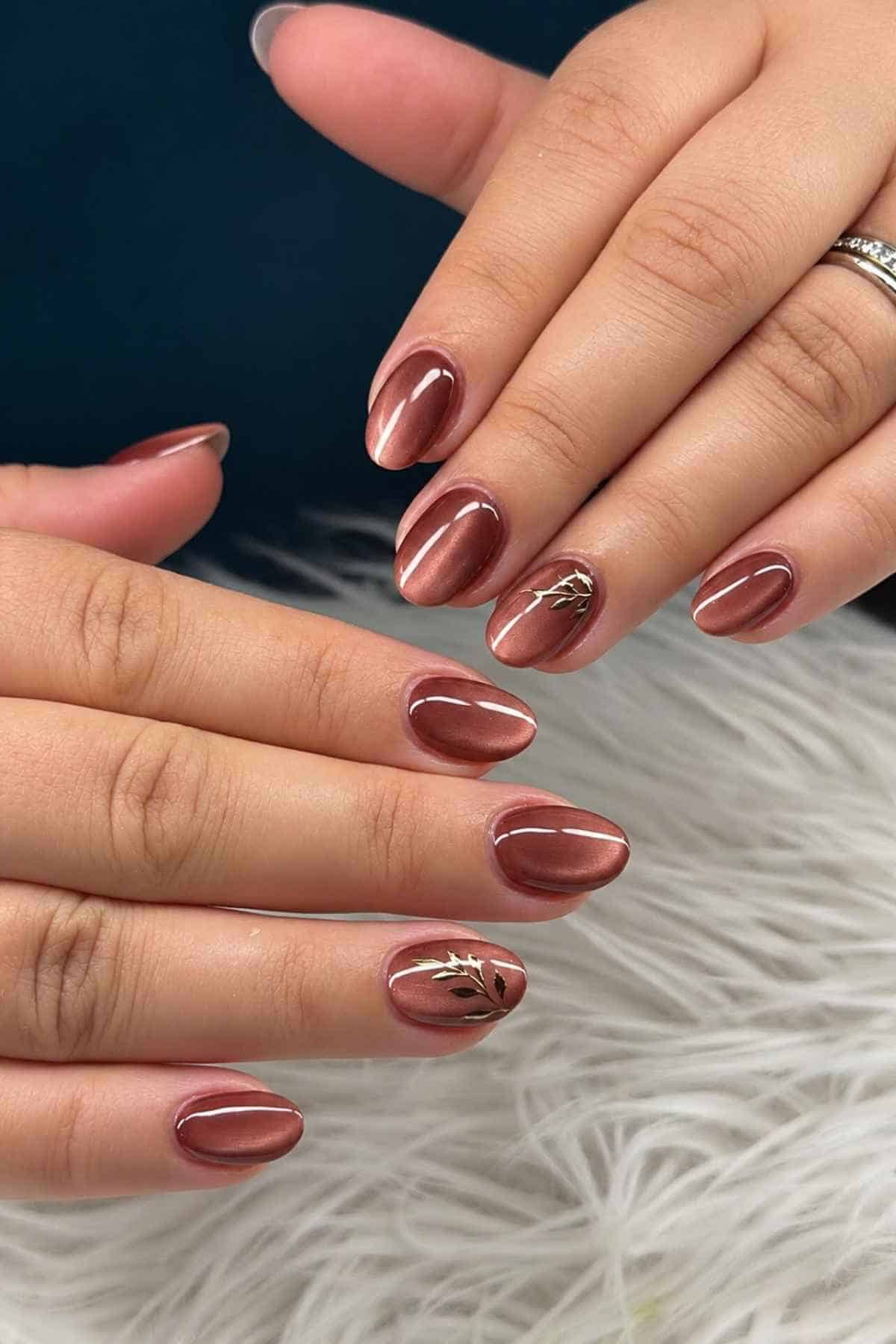 Hands with oval-shaped, metallic brown cat-eye nails, two of which feature small gold leaf accents.