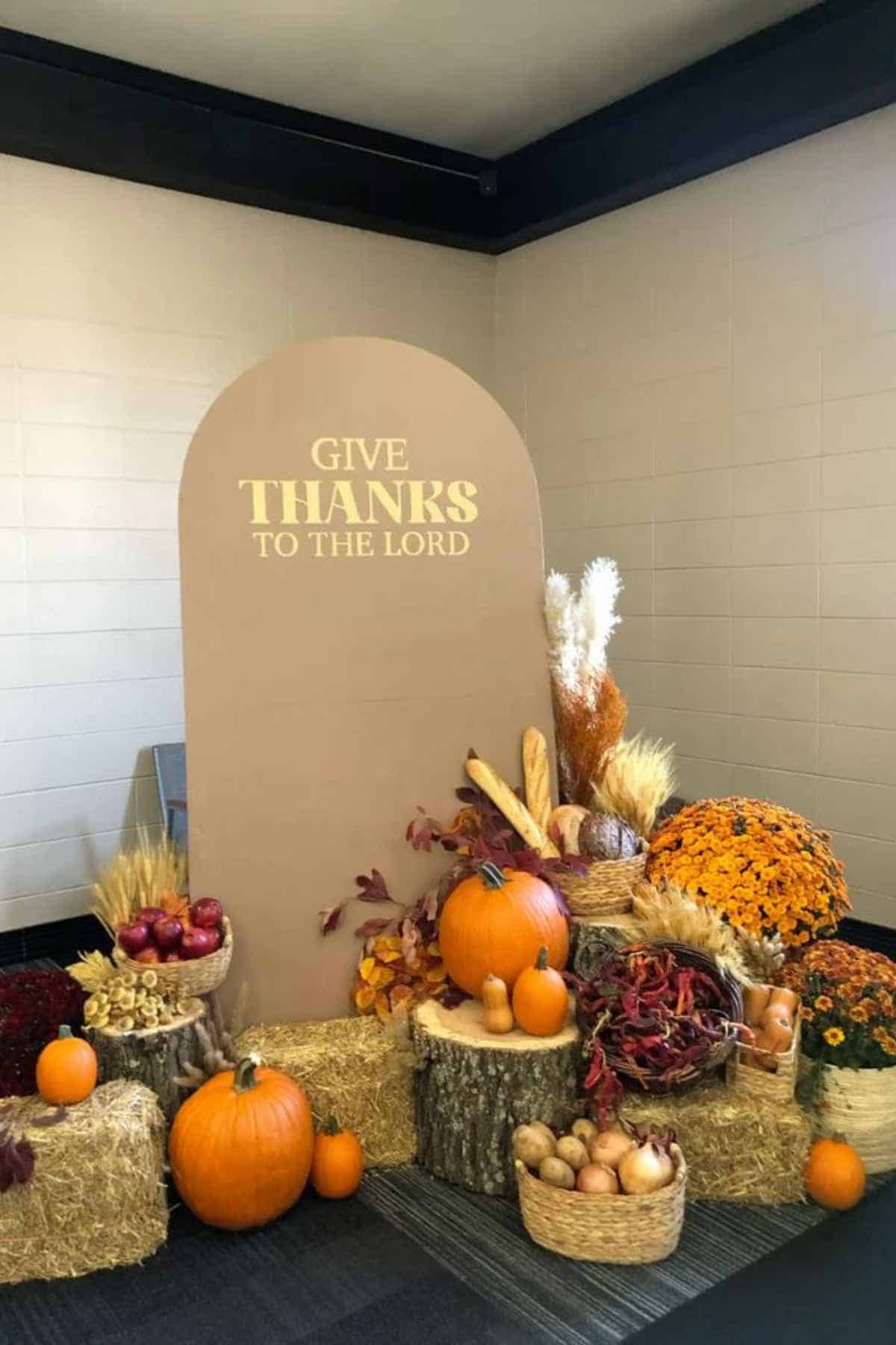 An arched sign that reads “GIVE THANKS TO THE LORD,” surrounded by pumpkins, gourds, dried wheat, flowers, and baskets of produce on hay bales and tree stumps.