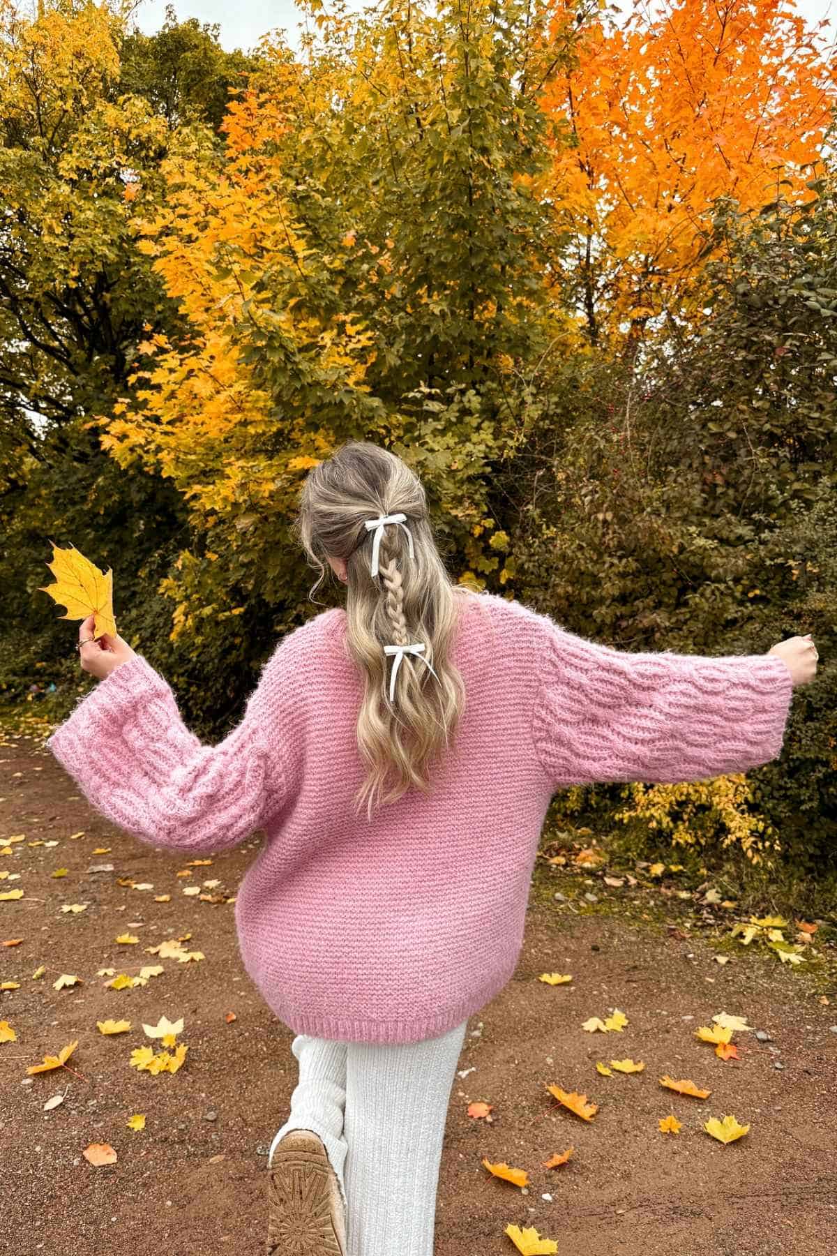 A person wearing a pink sweater with hair in a braided ponytail with white bows walks along a path lined with autumn trees, holding a yellow leaf.