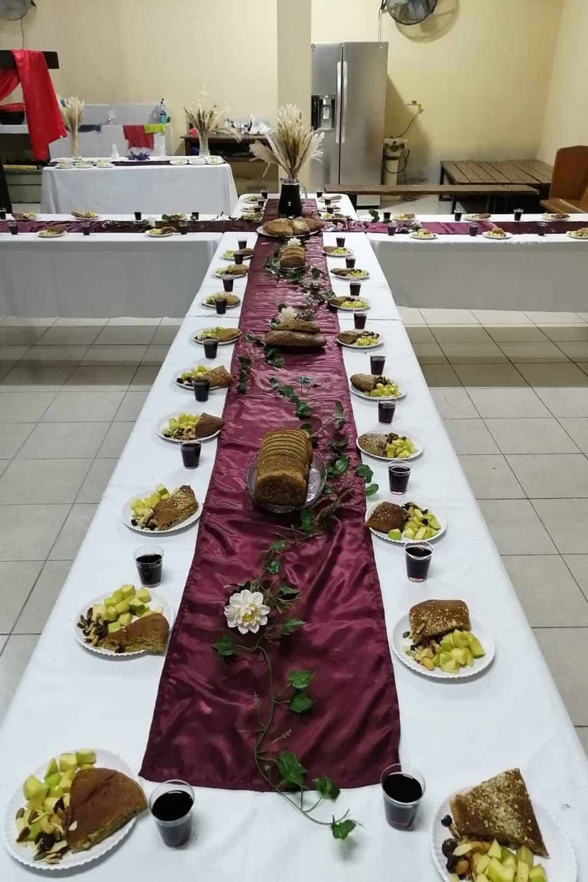 A long banquet table set with white tablecloths, a deep red runner, plates of sliced bread and fruit, and small cups of dark liquid.