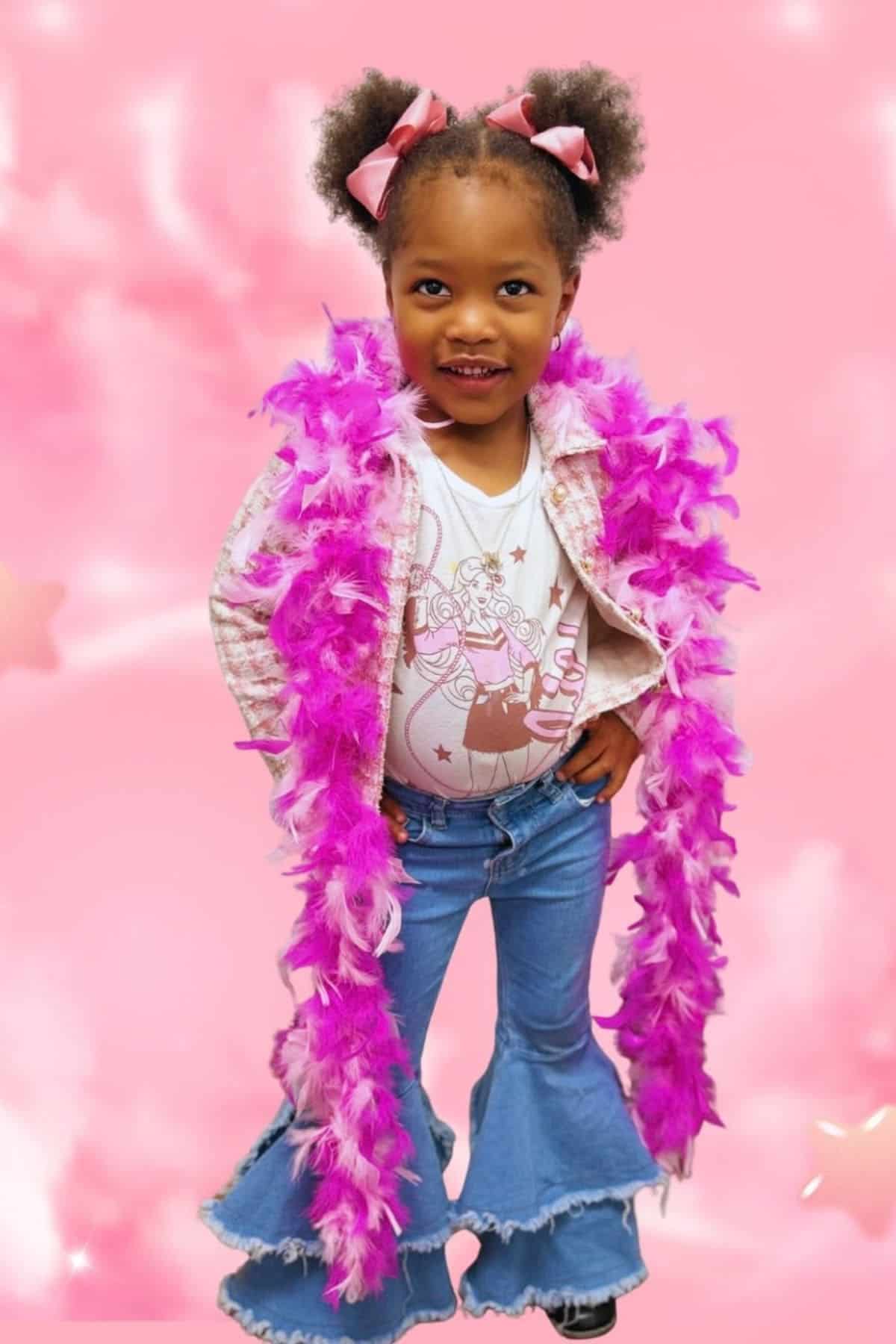 Young girl stands smiling in flared jeans, a graphic shirt, pink jacket, and a bright pink feather boa, with her hair in puffs accented by pink bows, against a pink background.