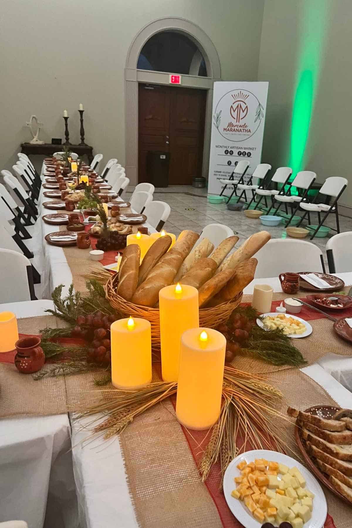 Long banquet table set for a meal with bread baskets, plates of cheese, and glowing LED candles, with a Mercado Maranatha sign in the background.