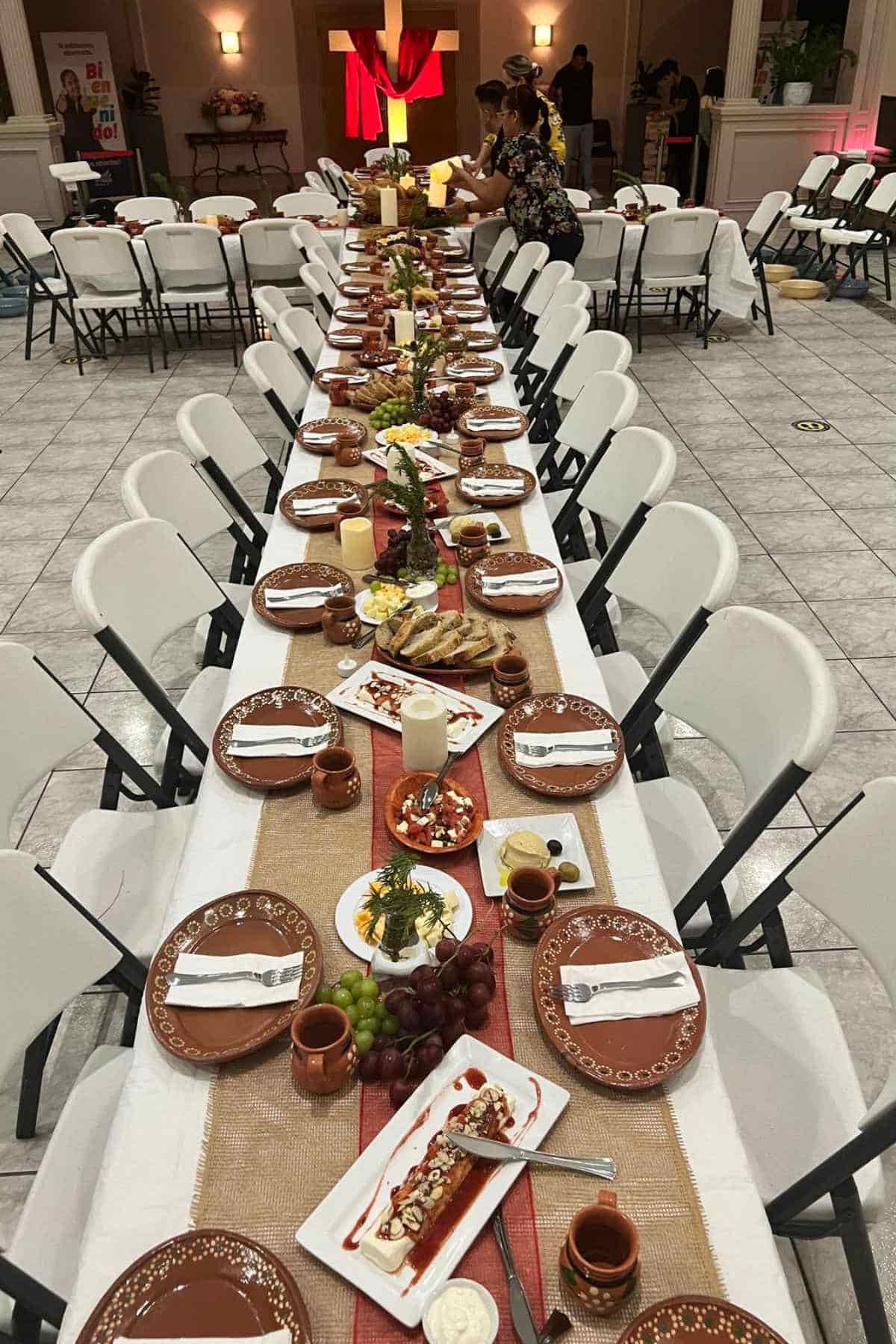 A long, rectangular table set for a formal meal with brown ceramic plates, silverware, and assorted dishes, surrounded by white folding chairs.