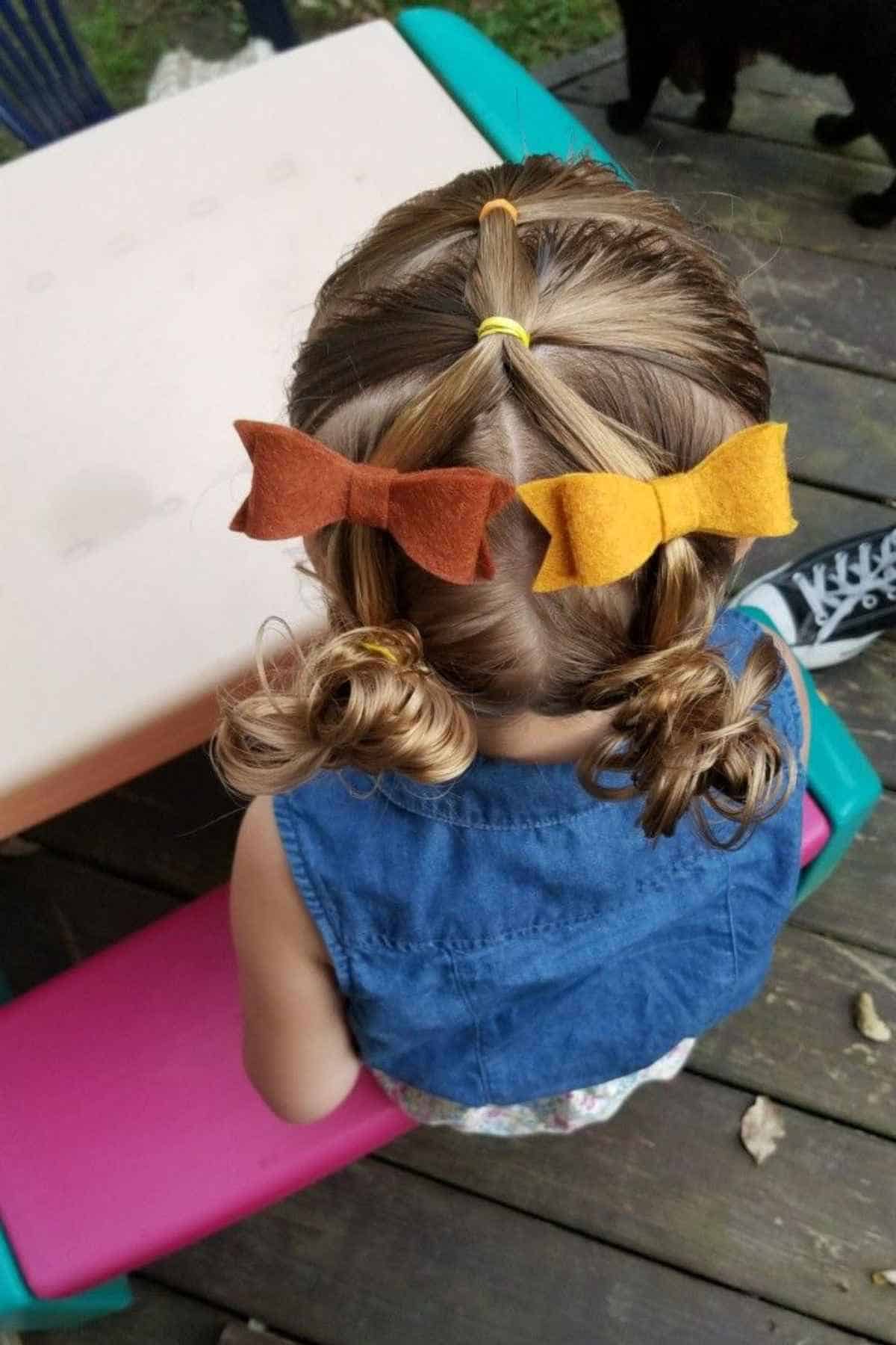 A child’s hair styled in small sections held by yellow elastics, accented with a brown felt bow and a mustard yellow felt bow above two small buns.