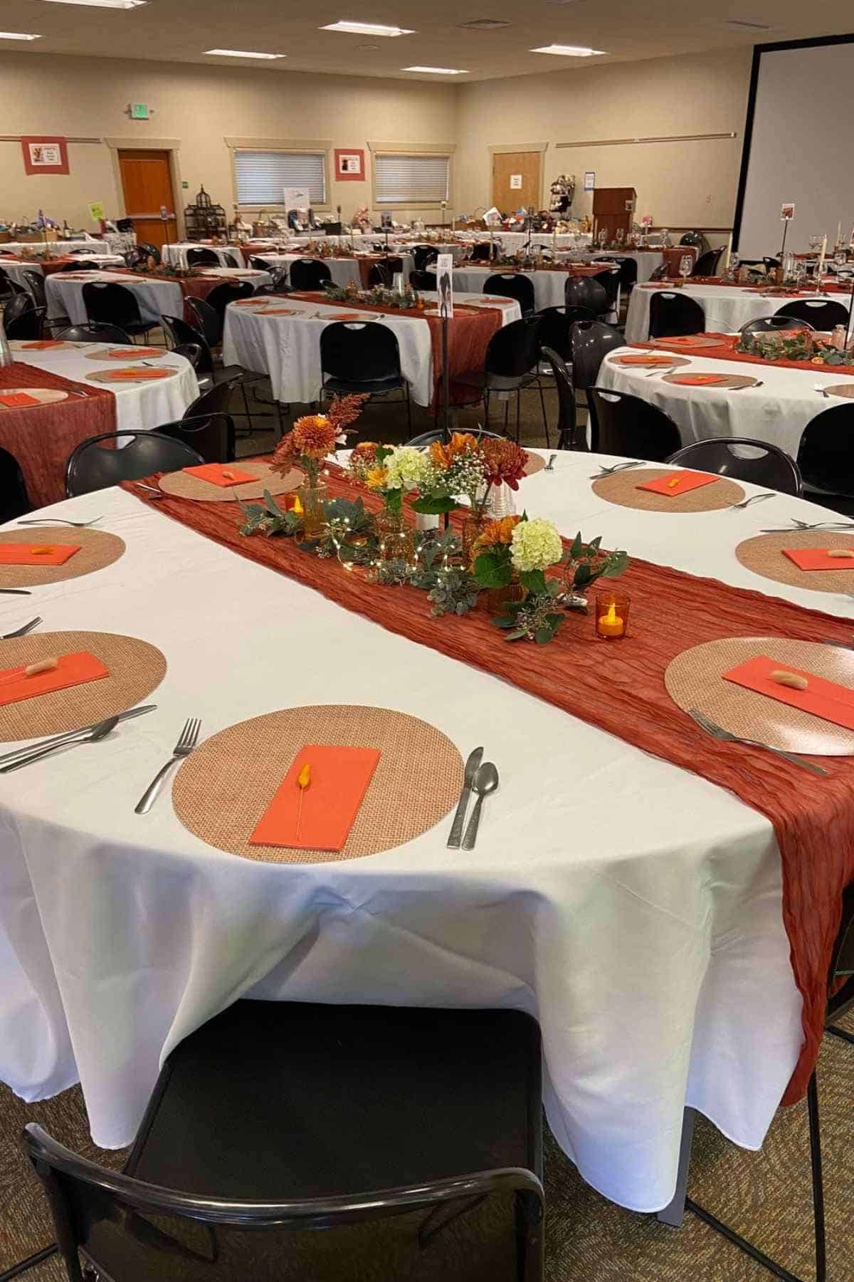 Round tables set for a banquet with white tablecloths, rust-colored runners, woven placemats, and floral centerpieces.