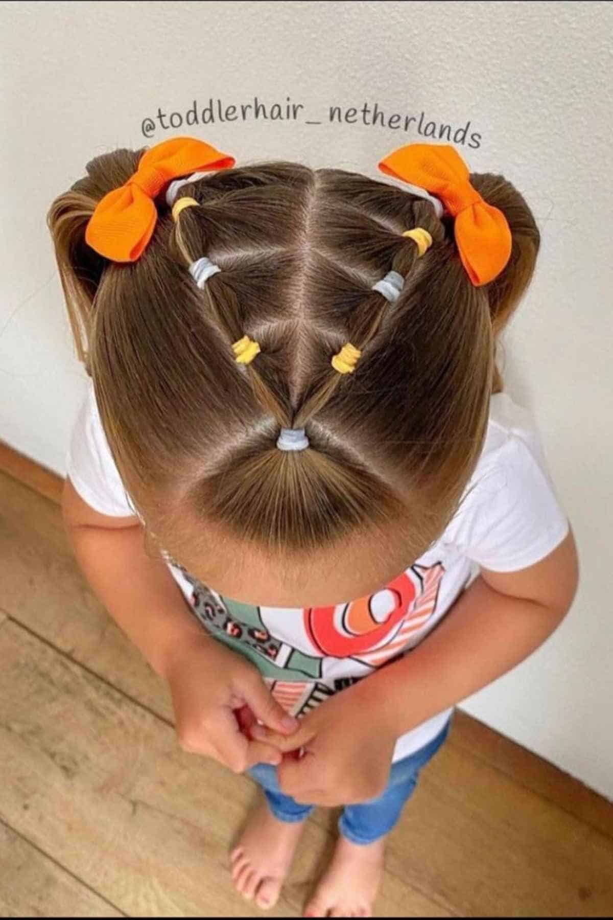 A child’s hair styled into pigtails with a crisscross pattern of colorful elastic bands and two orange bows.