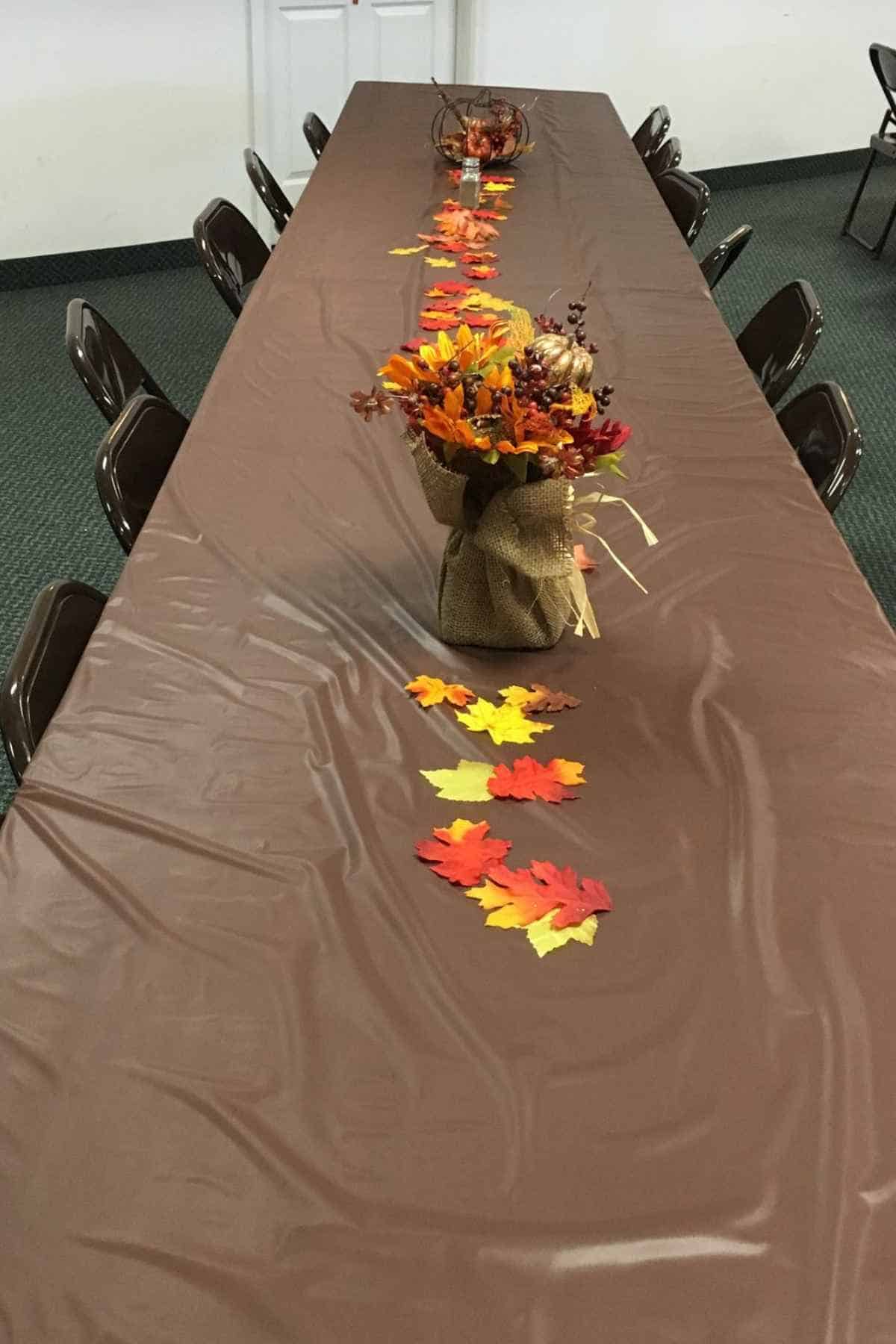 A long rectangular table covered in a brown cloth, decorated with a central autumn-themed floral arrangement and scattered faux fall leaves.