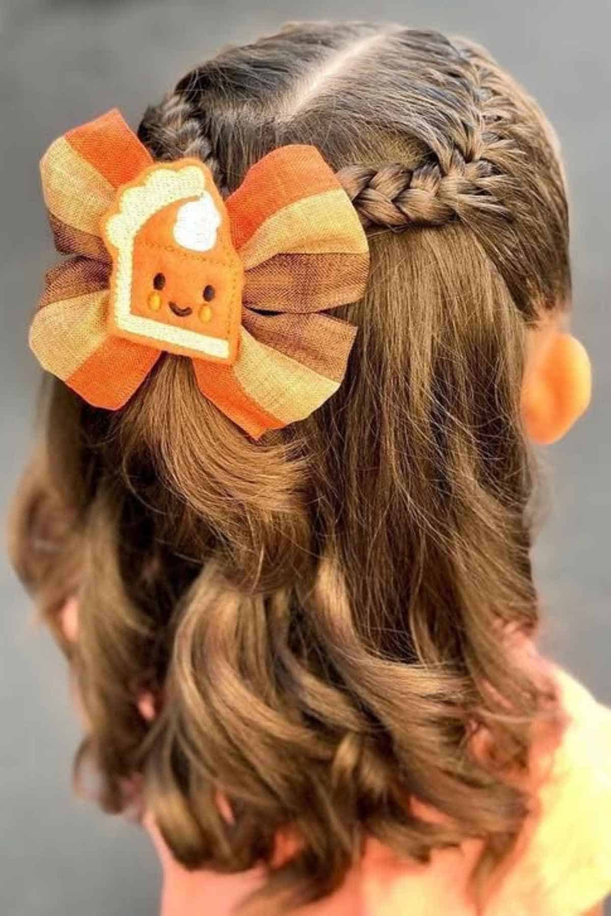 A person with braided, wavy hair wearing a hair bow featuring a cartoon pumpkin pie slice.